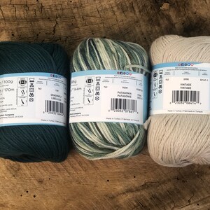 Op de afbeelding: Drie bollen garen in verschillende kleuren. De eerste bol is donkerblauwgroen, de tweede is een wit-groen gestreept garen en de derde is een lichtbeige garen. Elke bol heeft een etiket met de merknaam, de garennaam en de wasinstructies.