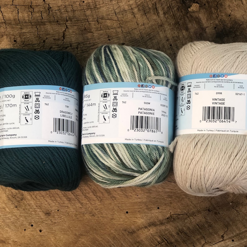 Op de afbeelding: Drie bollen garen in verschillende kleuren. De eerste bol is donkerblauwgroen, de tweede is een wit-groen gestreept garen en de derde is een lichtbeige garen. Elke bol heeft een etiket met de merknaam, de garennaam en de wasinstructies.