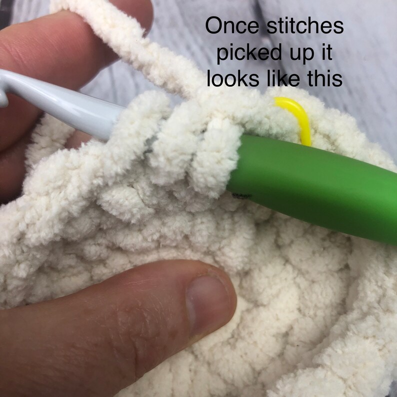 May include: A white plush yarn project in progress with a green crochet hook inserted into the stitches. A small yellow plastic stitch marker is attached to the work. The text "Once stitches picked up it looks like this" is visible.