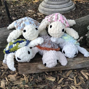 May include: A collection of four handmade crochet plush turtles featuring soft, chunky yarn in various colors. Each turtle has a white head with black button eyes and a uniquely patterned shell in shades of blue, pink, green, and brown.
