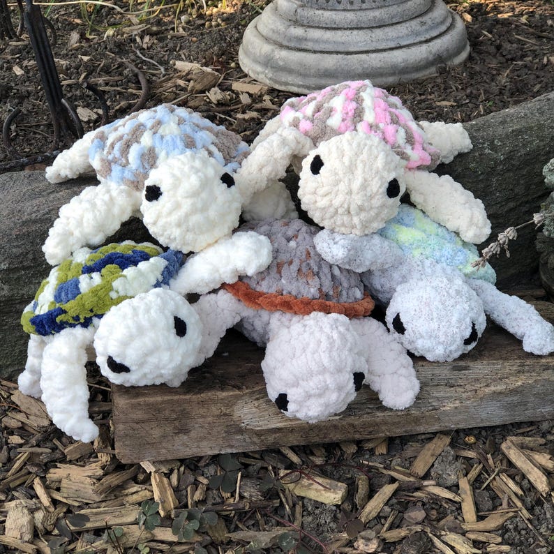 May include: A collection of four handmade crochet plush turtles featuring soft, chunky yarn in various colors. Each turtle has a white head with black button eyes and a uniquely patterned shell in shades of blue, pink, green, and brown.
