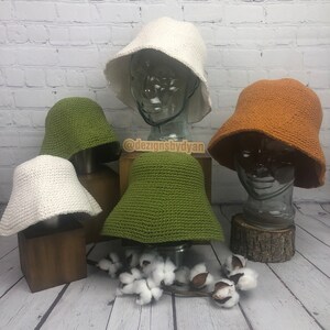 Peut inclure: Quatre chapeaux bob en crochet de différentes couleurs : blanc, vert, orange et vert foncé. Les chapeaux sont exposés sur des supports en bois et en verre. Les chapeaux sont tous réalisés avec un motif de point similaire.