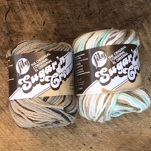 May include: Two skeins of Sugar'n Cream yarn in the colors "Salt" and "Driftwood". The yarn is a cotton blend and is made by Lily. The skeins are labeled "The Original L'Original El Original Sugar'n Cream".