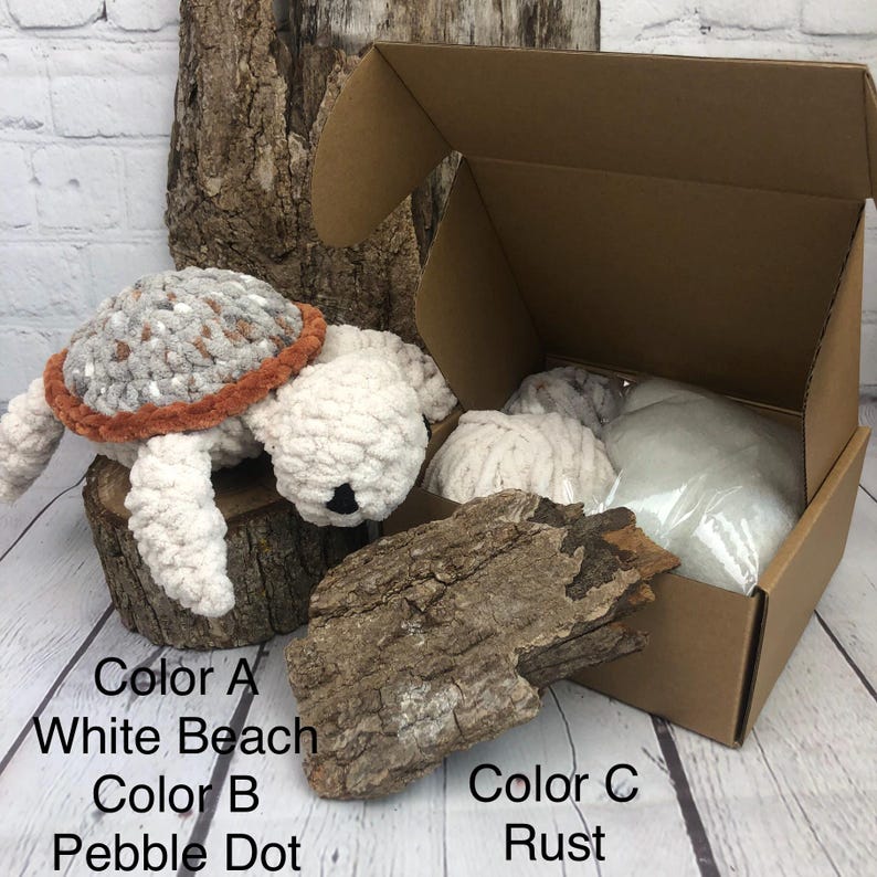 May include: A crochet turtle plush with a white body and a gray and white speckled shell with a rust-colored border. The set includes a brown cardboard box containing white yarn balls and a clear bag of stuffing, labeled Color A White Beach, Color B Pebble Dot, and Color C Rust.