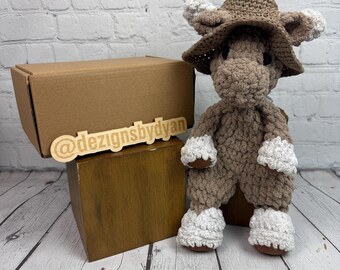 May include: A handmade crochet moose toy in shades of brown and white, wearing a matching hat. The toy is displayed on a wooden box with a cardboard box in the background. The wooden sign reads "@dezignsbydyan".