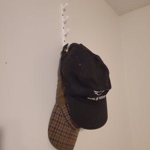 May include: A black and brown baseball cap with a white logo that says "MINI OF BEDFORD" hanging on a white wall mounted hook.
