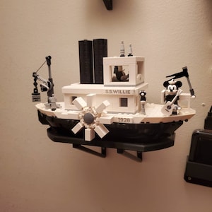 May include: A white and black LEGO model of the steamboat S.S. Willie, featuring a paddle wheel, a smokestack, and a figure of Mickey Mouse holding a guitar. The model has the text "S.S. WILLIE" and "1928" printed on it.
