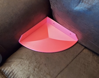 May include: A pink corner shelf with rounded edges and four mounting holes. The shelf is made of plastic and is designed to fit in the corner of a room.