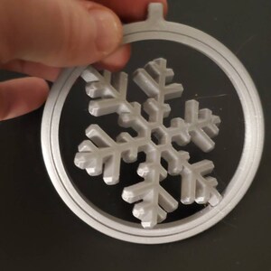 Könnte beinhalten: Ein weißer, 3D-gedruckter Schneeflocken-Anhänger mit einem runden Rahmen. Die Schneeflocke ist detailliert und filigran.