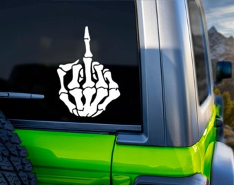 May include: A white skeleton hand giving the middle finger, a popular gesture of defiance, is displayed on a green car window.