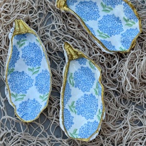 May include: Three oyster shells decorated with a blue hydrangea floral pattern on a white background. Each shell features a metallic gold painted edge along the rim. The shells are arranged in a cluster, showcasing the interior concave surface.