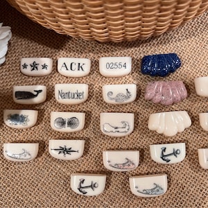 May include: A collection of 18 small, white ceramic tiles with various nautical designs. The tiles feature images of a whale, seashells, a compass, an anchor, a seahorse, a lighthouse, and the text "Nantucket".