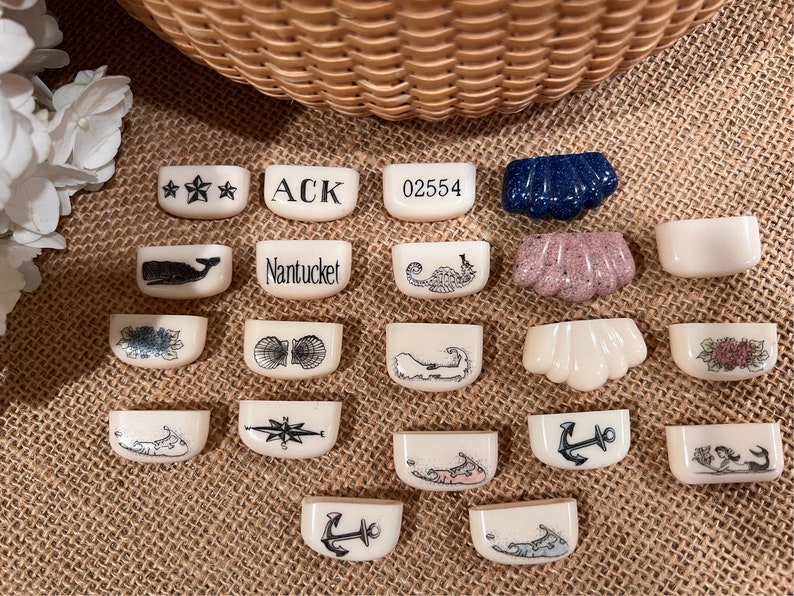 May include: A collection of 18 small, white ceramic tiles with various nautical designs. The tiles feature images of a whale, seashells, a compass, an anchor, a seahorse, a lighthouse, and the text "Nantucket".