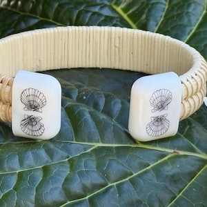 May include: Cream-colored woven bracelet with two rectangular, off-white clasps. Each clasp features a black seashell design. The bracelet is set against a backdrop of green leaves and a white flower.