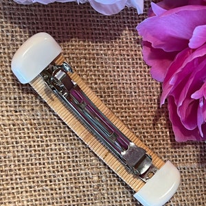 May include: A white and brown hair clip with a metal clasp. The clip is made of woven bamboo and has white plastic ends.