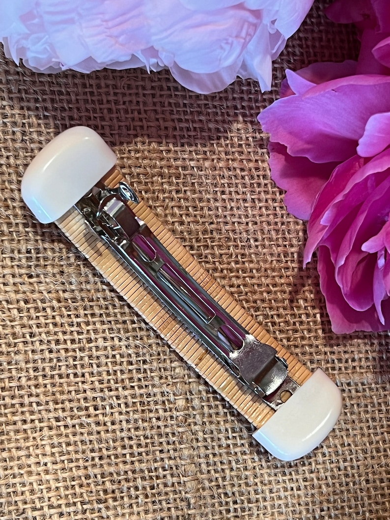 May include: A white and brown hair clip with a metal clasp. The clip is made of woven bamboo and has white plastic ends.