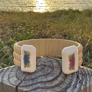May include: A beige woven bracelet with two rectangular ivory-colored end pieces. Each end piece features a floral design, one with blue and green flowers, the other with pink and green flowers. The bracelet is displayed on a wooden surface.