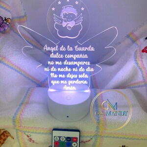 May include: A white acrylic night light with a colorful LED light base. The light features a design of an angel with wings and a halo, with the text "Angel de la Guarda. dulce compañía. no me desampares. ni de noche ni de dia No me dejes sola, que me perderia Amén" in Spanish.