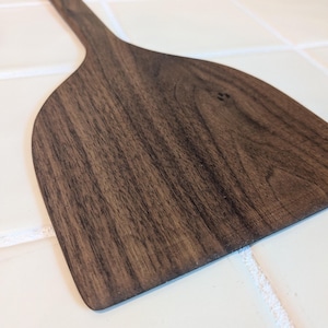 May include: A wooden spatula with a wide, curved head and a long, slender handle. The spatula is made of dark brown wood with visible grain patterns. The design is simple and functional, suitable for kitchen use.