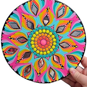May include: A colorful mandala painted on a round wooden surface. The mandala features a rainbow of colors, including pink, orange, yellow, green, blue, and purple. The center of the mandala is a bright pink circle surrounded by concentric circles of different colors. The mandala is decorated with small mirrors around the outer edge.