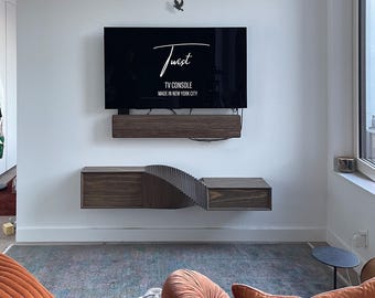 May include: A dark wood TV console with a unique wave-like design. The console is mounted on a white wall with a black TV above it. The TV screen displays the text "Twist TV Console Made in New York City".