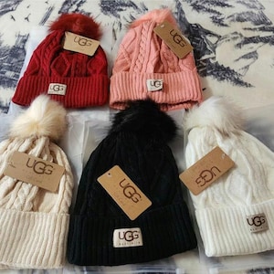 May include: Five cable-knit beanies in red, pink, black, cream, and white, each with a faux fur pom-pom. Each hat has a tag with the brand name "UGG" on it.