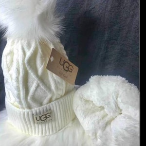 May include: White knitted beanie with a fluffy pom pom and a UGG Australia logo. The beanie is paired with a white fluffy ear warmer.