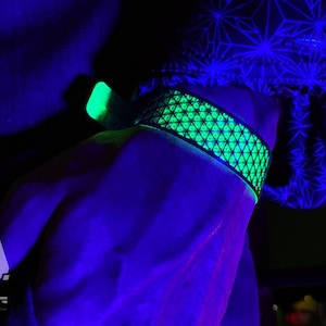 May include: A glowing green and black wristband with a geometric pattern. The wristband is illuminated under a black light, showcasing a vibrant neon green color. The image is taken in a dark environment, highlighting the glowing effect.
