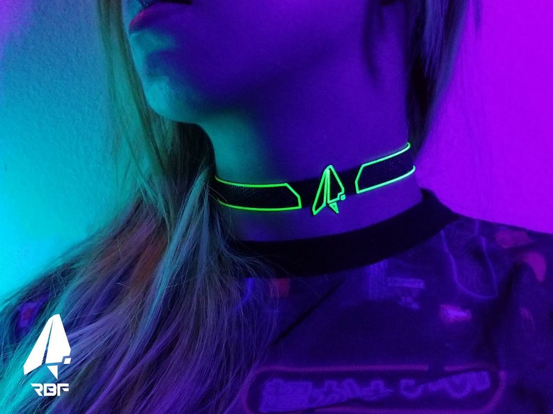 K&ouml;nnte beinhalten: Schwarze Halskette mit neongr&uuml;nen Zierleisten und einem geometrischen Design. Die Halskette leuchtet neongr&uuml;n und bildet einen Kontrast zum schwarzen Material. Das RBF-Logo ist unten links sichtbar.