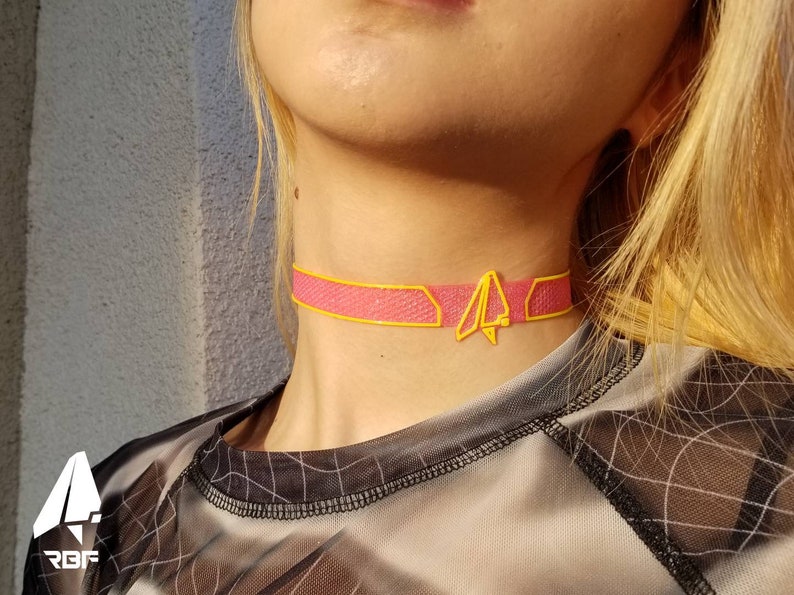 K&ouml;nnte beinhalten: Halsband in Rosa mit gelbem Besatz und geometrischem Design. Die Halskette wird um den Hals getragen. Der Choker hat eine gelbe geometrische Form in der Mitte. Die Halskette besteht aus einem Mesh-Material.