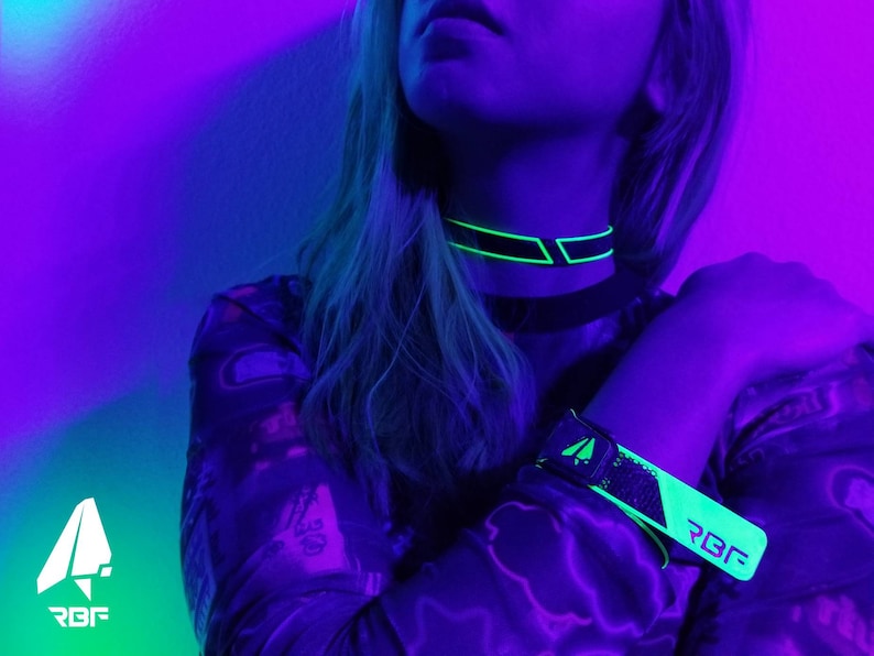 K&ouml;nnte beinhalten: Ein schwarzes Choker- und Armband-Set mit neongr&uuml;nen Akzenten. Das Choker und Armband haben ein geometrisches Design mit den Buchstaben "RBF" sichtbar. Das Bild wird von violetten und blauen Neonlichtern beleuchtet.