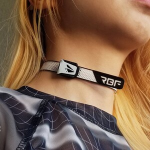 K&ouml;nnte beinhalten: Ein wei&szlig;-schwarzes Choker-Halsband mit den Buchstaben "RBF" darauf. Das Halsband hat ein Mesh-Design und einen silbernen Verschluss. Die Person auf dem Bild tr&auml;gt ein schwarz-wei&szlig; gemustertes Oberteil.