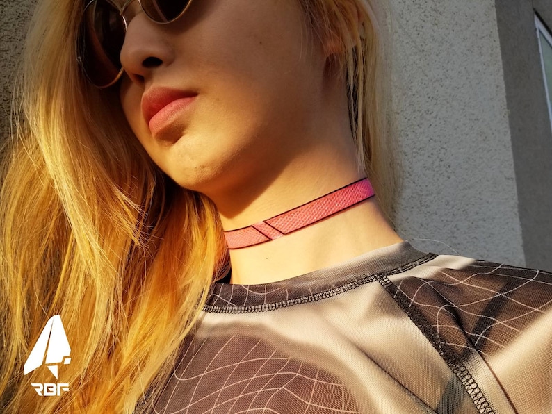 K&ouml;nnte beinhalten: Ein pinkfarbenes Choker-Halsband mit geometrischen Akzenten. Das Halsband besteht aus einem strukturierten Material und wird um den Hals getragen. Das Bild zeigt auch eine Person, die ein Mesh-Top und eine Sonnenbrille tr&auml;gt.