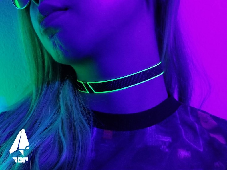 K&ouml;nnte beinhalten: Schwarze Halskette mit neongr&uuml;nen geometrischen Akzenten. Die Halskette wird um den Hals getragen und vor einem Hintergrund aus lila und gr&uuml;ner Beleuchtung platziert. Das RBF-Logo ist unten links sichtbar.