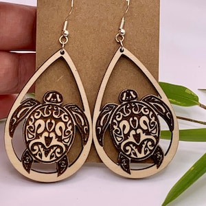 May include: A pair of wooden teardrop earrings with a laser-cut sea turtle design. The earrings are light brown with a detailed dark brown turtle carving. The earrings hang from silver-colored fish hook ear wires.