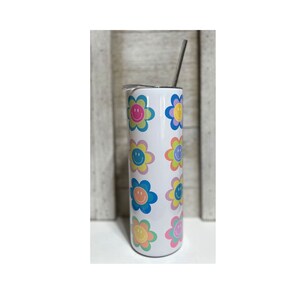 Pu&ograve; includere: Un bicchiere bianco con un motivo ripetuto di disegni floreali colorati, ognuno con una faccia sorridente al centro. I fiori hanno petali rosa pastello, blu, gialli e verdi. Una cannuccia argentata si estende dalla parte superiore.