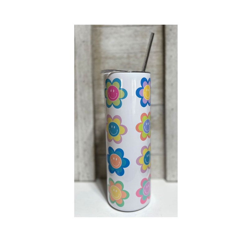 Pu&ograve; includere: Un bicchiere bianco con un motivo ripetuto di disegni floreali colorati, ognuno con una faccia sorridente al centro. I fiori hanno petali rosa pastello, blu, gialli e verdi. Una cannuccia argentata si estende dalla parte superiore.