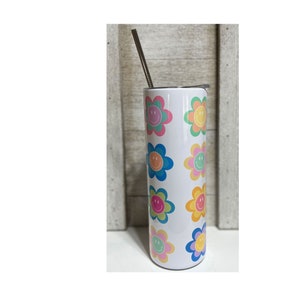 Pu&ograve; includere: Un bicchiere bianco con un motivo ripetuto di fiori sorridenti colorati. I fiori hanno petali rosa, blu, gialli e verdi con una faccia sorridente al centro. Una cannuccia d'argento &egrave; inserita nel coperchio.