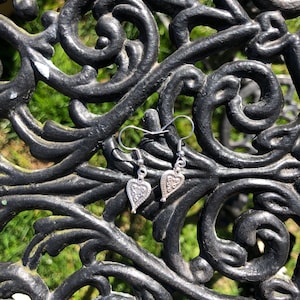 May include: A pair of silver heart-shaped earrings with intricate patterns. The earrings are suspended from silver hooks and rest on a black, ornate metal surface with a green, blurred background.