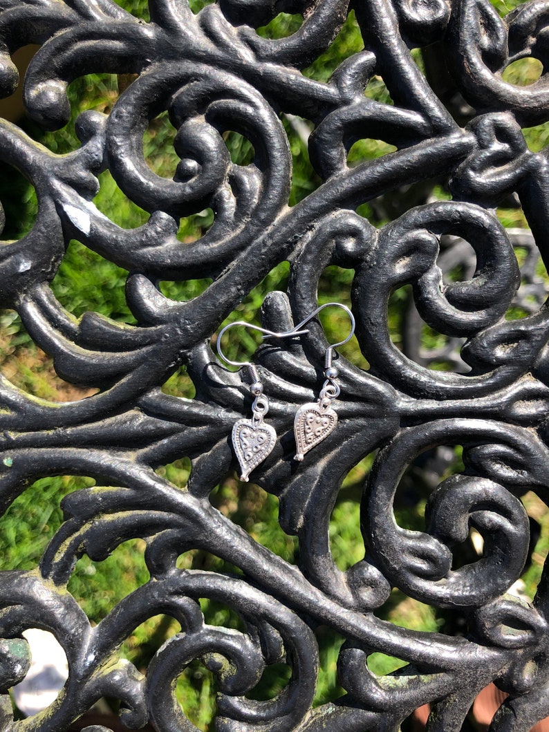 May include: A pair of silver heart-shaped earrings with intricate patterns. The earrings are suspended from silver hooks and rest on a black, ornate metal surface with a green, blurred background.