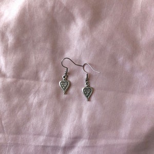 May include: A pair of silver-colored heart-shaped earrings. Each earring features a small heart pendant with a textured design. The earrings hang from a simple hook. The background is a light pink fabric.