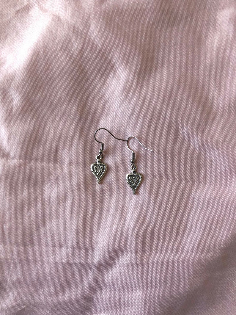 May include: A pair of silver-colored heart-shaped earrings. Each earring features a small heart pendant with a textured design. The earrings hang from a simple hook. The background is a light pink fabric.