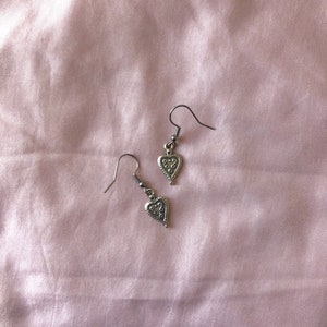 May include: A pair of silver-colored heart-shaped earrings with intricate detailing. The earrings hang from a simple hook design. The background is a soft, pink fabric.