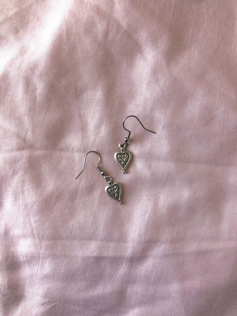May include: A pair of silver-colored heart-shaped earrings with intricate detailing. The earrings hang from a simple hook design. The background is a soft, pink fabric.
