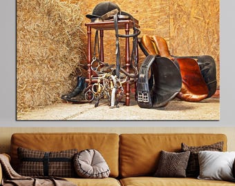 May include: A brown wooden stool holds a black riding helmet and leather bridle with metal bits. A brown and black leather saddle rests beside the stool, with a black riding boot nearby, all positioned against a backdrop of a large hay bale.