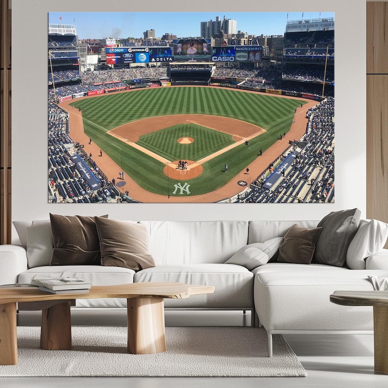 May include: An aerial view of a baseball field with green grass and a brown dirt infield. The field is surrounded by a large stadium with bleachers filled with fans. The New York Yankees logo is visible in the center of the field.