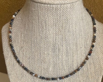 May include: A beaded necklace displayed on a mannequin. The necklace features small, round beads in shades of brown, gray, white, and blue. The beads are strung together to form a delicate necklace. The mannequin is covered in a beige fabric.