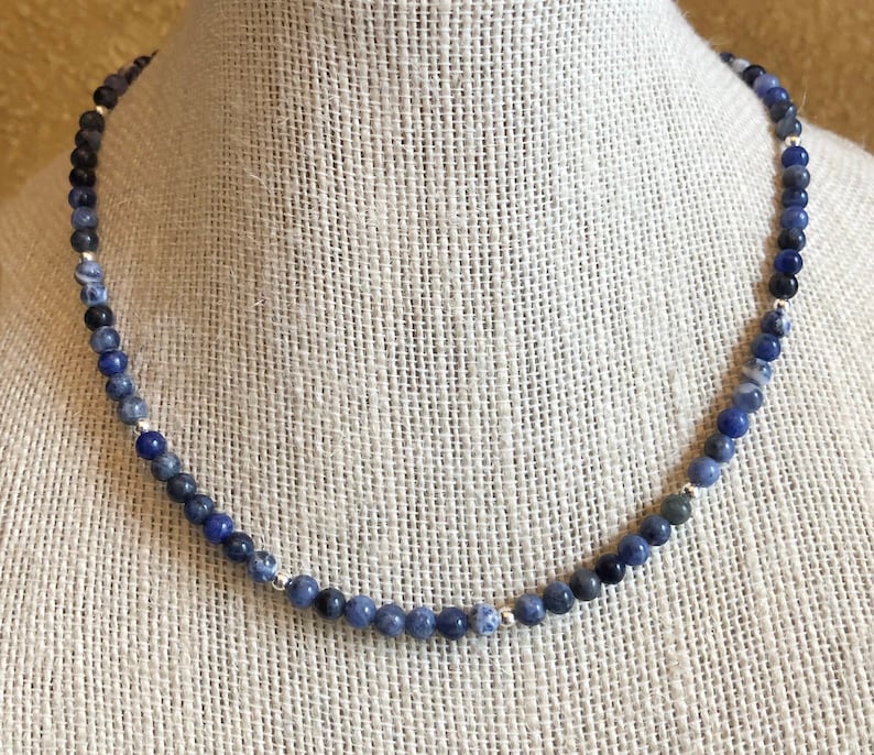 May include: A blue and black gemstone necklace with silver beads. The necklace is made of round beads and is displayed on a fabric-covered mannequin.