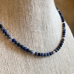 May include: A blue and black gemstone necklace with silver beads. The necklace is made of small, round beads that are strung together on a silver chain.