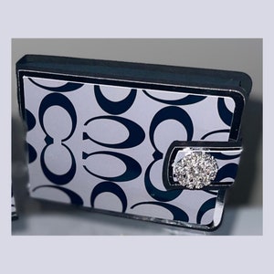 May include: A small, rectangular wallet with a white background and a navy blue repeating pattern. The wallet has a black border and a silver clasp with a cluster of clear rhinestones.
